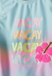 Világos kék fürdőruha, amely multiszínű csillámos felirattal rendelkezik, ami "VACAY" feliratot formál pink, narancssárga, zöld és kék színekben, egy pink flitteres virágdíszítéssel.