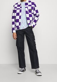 Lila och vit rutigt jacka med en kordfläta krage, lager på lager över en tie-dye t-shirt; matchat med mörka cargobyxor och svarta sneakers.