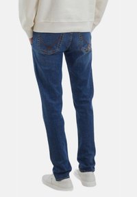 Jeans in denim blu con vestibilità dritta, caratterizzati da un lavaggio delicato, tasche posteriori con cuciture diagonali, indossati con sneakers bianche.