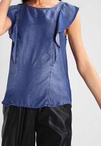 Denim top in dark blue with short ruffled sleeves, round neckline, and subtle seam detailing, paired with faux leather pants.