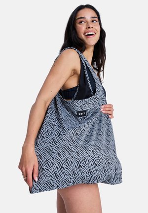 Joven sonriente, llevando un bolso grande Roxy a rayas de cebra en negro y gris sobre el hombro, con camiseta negra.