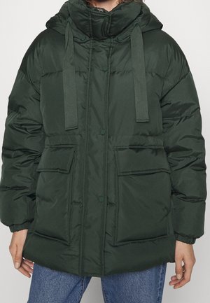 Veste matelassée à capuche vert foncé avec grandes poches avant et fermeture à boutons portée sur un jean bleu