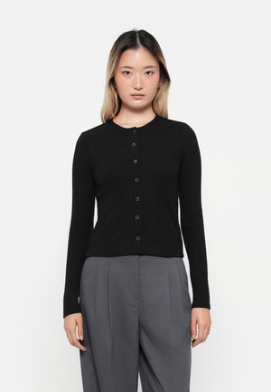 Black ribbed cardigan with long sleeves and round neckline, featuring a button-down front. Worn over light gray high-waisted trousers.