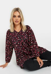 Svart blommig blus med långa ärmar, V-ringning och en lös passform med röda och krämfärgade blommor mot en mörk bakgrund.