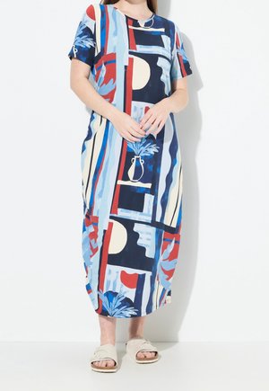 Femme portant une robe midi à motif abstrait coloré avec des manches courtes et des sandales beiges à enfiler, debout devant un fond blanc.