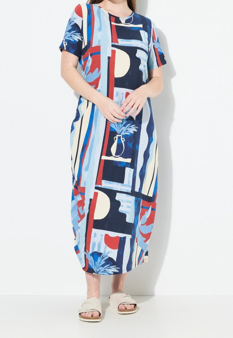 Femme portant une robe midi à motif abstrait coloré avec des manches courtes et des sandales beiges à enfiler, debout devant un fond blanc.