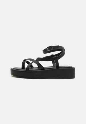 Zwarte platform sandalen met brede kruisbanden, een enkelband met een gesp en een gladde matte afwerking. De zool is dik en gestructureerd.