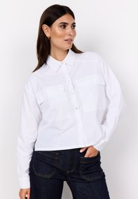 White button-up shirt with a collar, long sleeves, and two chest pockets. Material is smooth and lightweight; cropped design.