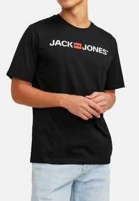 Schwarzes Baumwoll-T-Shirt mit einem weißen Logo "JACK & JONES" und einem roten rechteckigen Akzent. Kurze Ärmel und runder Halsausschnitt. Lässige Passform.