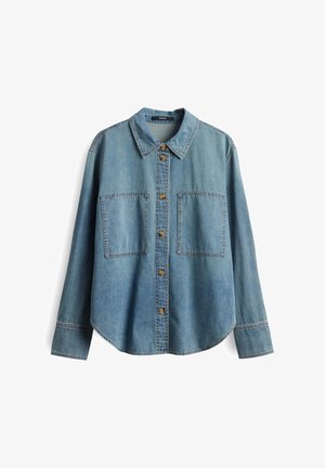 Chemise en denim bleu clair avec un col pointu, des boutons en bronze, deux grandes poches poitrine et des manches longues. Présente un ourlet arrondi.