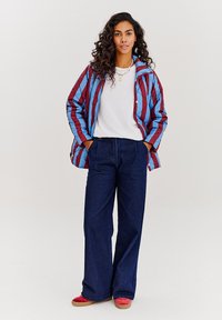 Prošívaná bunda v modro-vínových pruzích, bílý tričko, tmavě modré široké džíny a červené boty. Kadeřavé vlasy dodávají outfitu texturu.