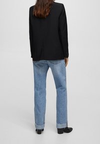 Svart blazer med strukturerade axlar, slät textur och knäppta ärmar, parat med ljusblå raka jeans och svarta klackstövlar.