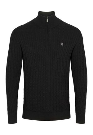 Un homme souriant porte un pull noir en maille torsadée à demi-zip sur une chemise blanche et un jean foncé, sur un fond uni et clair.