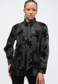 Čierna priehľadná blúzka s kvetinovým vzorom, dlhými rukávmi, golierom na gombíky a predným viazaním. Textúrovaná látka s hladkým povrchom.