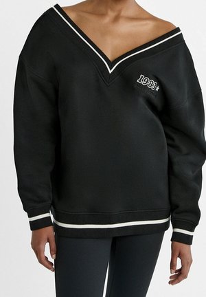 Zwarte oversized V-hals trui met witte bies aan kraag, manchetten en zoom, met de tekst "1983 Manhattan" op de linkerborst, gedragen door een persoon.