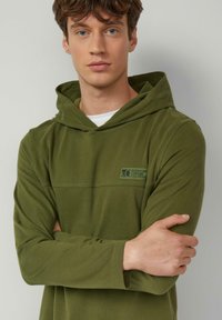 Olijfgroene hoodie van zachte, lichte stof met een logopatch aan de voorkant en stiksels langs de naden. Voorzien van een ruime capuchon.