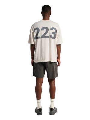 Man staat met zijn rug naar de kijker, draagt een beige oversized T-shirt met "223" in grote grijze cijfers, donkere korte broek, witte sokken en zwarte sneakers.