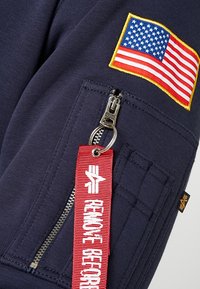 Felpa blu navy con una toppa della bandiera americana e un'etichetta rossa "RIMUOVERE PRIMA DEL VOLO", dotata di una tasca con zip e tessuto testurizzato.