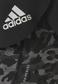 Yksityiskohta mustasta ja harmaasta camouflagemuotoisesta Adidas Primeblue-kankaasta, jossa näkyvissä vetoketju ja valkoinen Adidas-logo.