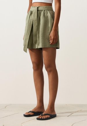 Nő, aki olíva zöld magasderekú rövidnadrágot visel anyagövet, fehér crop topot és fekete flip-flop papucsot, világos színű padlólapokon áll.