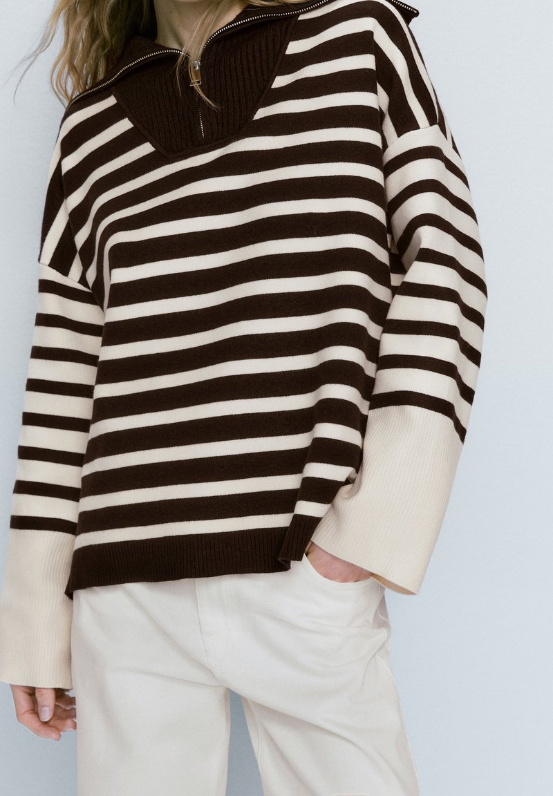 Pull rayé marron et crème avec un col haut et une fermeture éclair, présentant une coupe ample et des poignets côtelés. Associé à un pantalon de couleur claire.