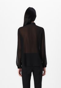 Blusa preta transparente com mangas longas, punhos abotoados e colarinho. Tecido fluido com um corte solto, combinada com calças pretas.