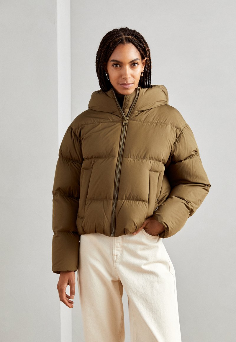 Une veste en duvet marron avec un col haut et une fermeture éclair, ayant une forme carrée, une texture matelassée et de grandes poches latérales.