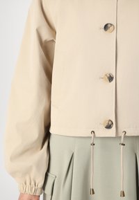 Beige cropped jasje met opgeblazen mouwen, knoopsluiting en trekkoorden in de zoom. Draagt over een lichtgroene geplooide onderkant.