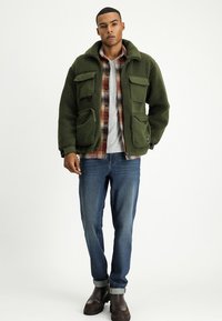 Zielona polarowa kurtka z dwoma przednimi kieszeniami, założona na koszulę w kratę, w połączeniu z niebieskimi dżinsami i brązowymi butami. Prosty, teksturowany wzór.
