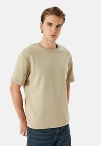 T-shirt in cotone beige con collo a giro e maniche corte, caratterizzata da una vestibilità rilassata e una texture liscia. Indossata con jeans in denim blu.