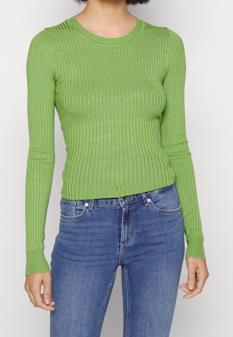 Femme portant un pull vert côtelé ajusté à manches longues, rentré dans un jean bleu de lavage moyen, debout devant un fond uni.