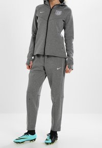 Chándal gris de Nike con capucha con cremallera y pantalones ajustados. Presenta un emblema bordado y un logotipo blanco en contraste. Zapatillas negras y verde menta.