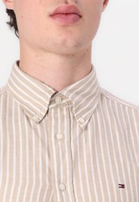 Camisa de botones a rayas beige y blanca con un cuello clásico y un pequeño logotipo en el lado izquierdo del pecho. Tela de algodón con un acabado texturizado.