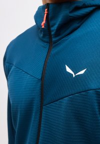 Μπλε μπουφάν με φερμουάρ και ψηλό γιακά, μαύρο φερμουάρ, κόκκινη ρυθμιζόμενη κορδόνι και λευκό στυλιζαρισμένο λογότυπο πουλιού στο στήθος.