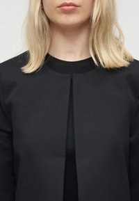 Femme blonde portant un haut noir à col rond et une veste noire ouverte sur le devant, montrée des épaules aux lèvres sur un fond neutre.