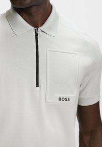 Man wearing a light gray polo shirt with a black zipper and a raised rectangular design reading "BOSS" on the chest.