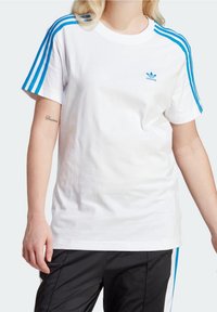 Vit bomull t-shirt med blå tre-rands accenter på axlarna och liten blå Adidas-logotyp på bröstet. Kort ärm, avslappnad passform.