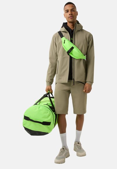 Beige waterproof jacket over black top, green fanny pack, khaki shorts, and grey sneakers. Holding a large green duffel bag with black accents.
