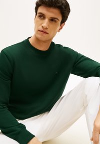 Σκούρο πράσινο βαμβακερό φούτερ με στρογγυλή λαιμόκοψη, μανσέτες ριμπ και μικρό λογότυπο στο στήθος. Συνδυάζεται με λευκό παντελόνι.