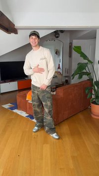 Ung mann som smiler med øynene lukket, iført kamuflasjebukser, beige genser og caps, stående i en moderne stue med oransje sofa og stor plante.