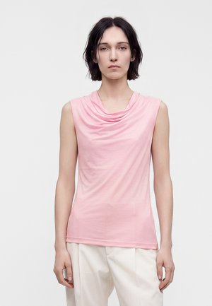 Vrouw met kort donker haar, die een mouwloze roze top en witte broek draagt, staand, voorover gericht tegen een effen lichte achtergrond.