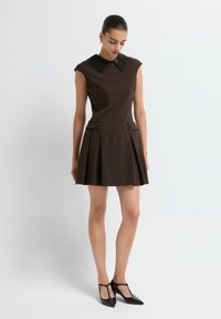 Braunes ärmelloses Kleid mit einem strukturierten schwarzen Kragen, faltigem Rock und dekorativen Knöpfen an der Taille. Getragen mit schwarzen spitzen Schuhen.