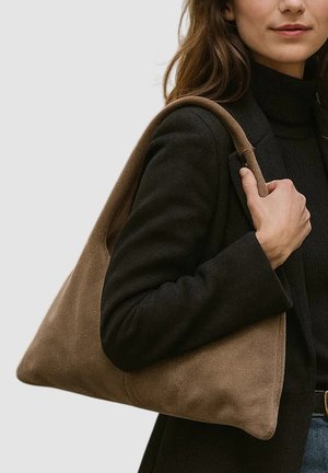 Sac bandoulière en daim marron clair avec une forme triangulaire, une texture lisse et une large sangle d'épaule, porté par une personne vêtue de vêtements foncés.