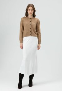 Bruine gebreide cardigan met ronde hals en knoopsluiting, gecombineerd met een witte midirok en kniehoge zwarte laarzen. Soepele texturen.