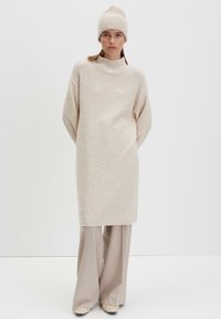 Abito in maglia beige oversize con collo alto, abbinato a pantaloni larghi beige e un berretto a coste coordiato. Texture liscia, design minimalista.