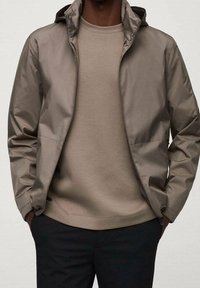 Hellbraune, wasserabweisende Jacke mit Kapuze, Reißverschluss vorne und Seitentaschen, über einem passenden Rundhalsausschnitt-Sweatshirt getragen.