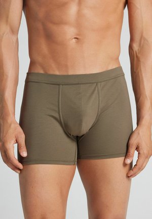 Männlicher Oberkörper trägt olivgrüne enganliegende Boxer-Shorts, steht mit locker an den Seiten hängenden Armen vor einem schlichten hellen Hintergrund.