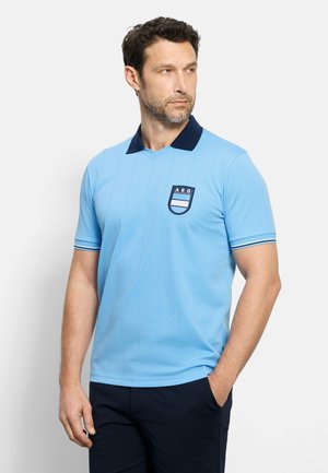 Man draagt lichtblauw poloshirt met donkere kraag en ARG-wapen, staat met één hand in de zak, kijkt naar rechts op een effen achtergrond.