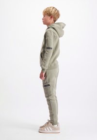 Lichtgroene katoenen hoodie met een voorzak, ribgebreide boorden en een zwart patch op de mouw. Gecombineerd met taps toelopende trainingsbroek en beige sneakers.