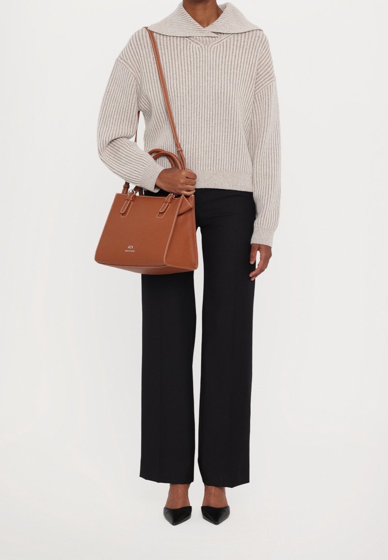 Personne portant un pull beige côtelé, un pantalon noir à jambes larges, des chaussures noires pointues, tenant un sac à main en cuir marron avec bandoulière.